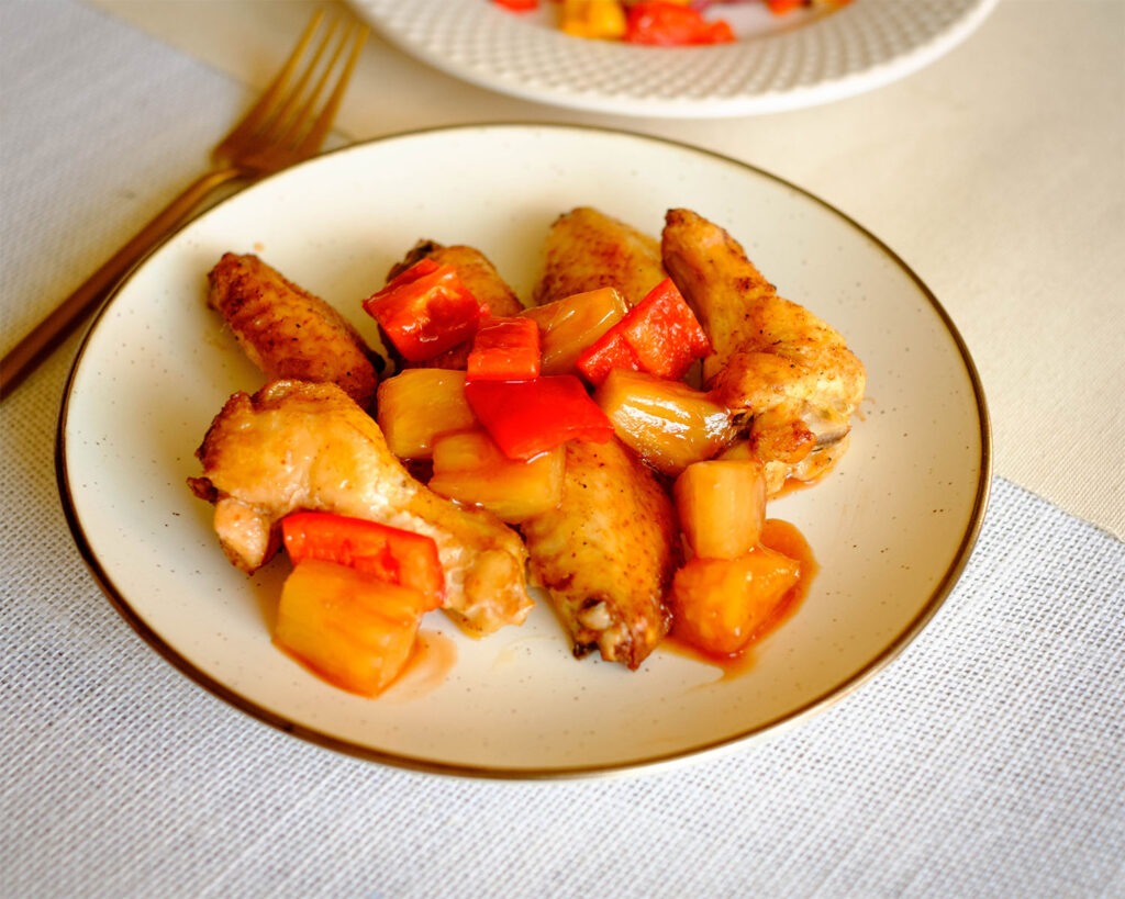 Round plate with chicken wings drizzled with sweet and sour sauce that includes pineapple chunks and red bell peppers.
