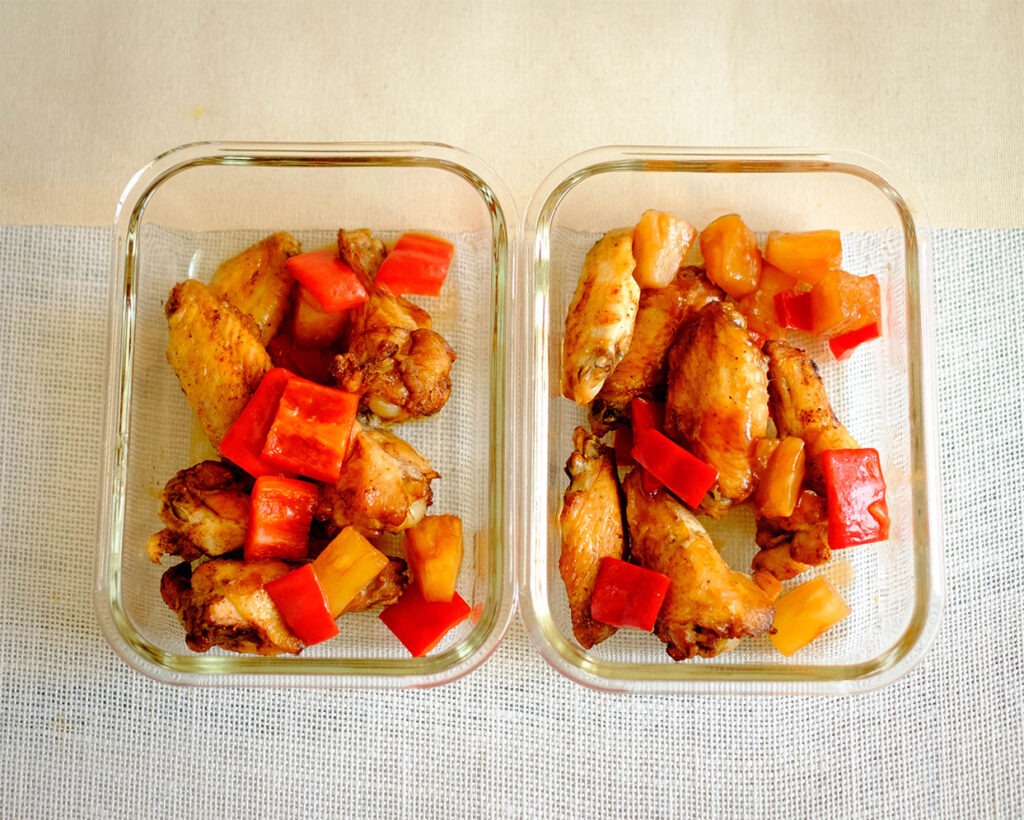 Glass meal prep container with chicken wings drizzled with sweet and sour sauce that includes pineapple chunks and red bell peppers.