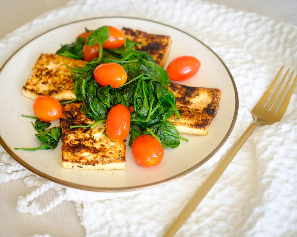 Round plate with pan-fried tofu topped with sauteed spinach and cherry tomatoes.
