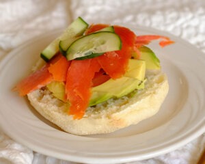 White round plate with open faced english muffin topped with sliced avocado, smoked salmon, and sliced cucumber.