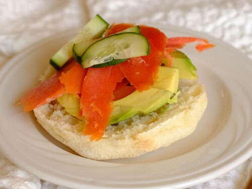White round plate with open faced english muffin topped with sliced avocado, smoked salmon, and sliced cucumber.