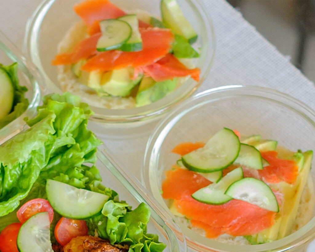Round glass meal prep containers with open faced english muffin topped with sliced avocado, smoked salmon, and sliced cucumber.
