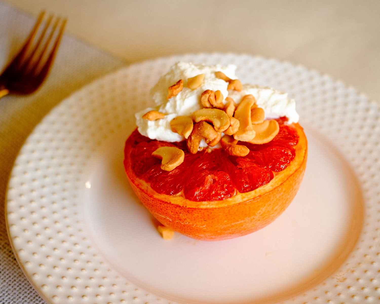 Round white plate with a grapefruit half, topped with yogurt and cashew nuts.