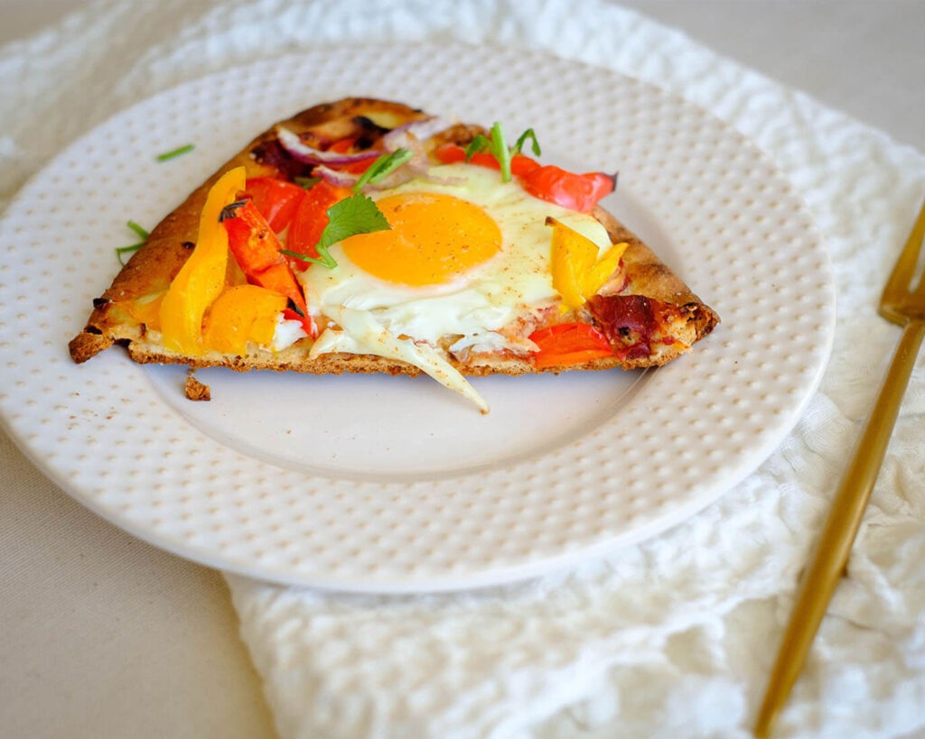 Round plate with breakfast pizza that has egg topping, bell pepper, and red onions.