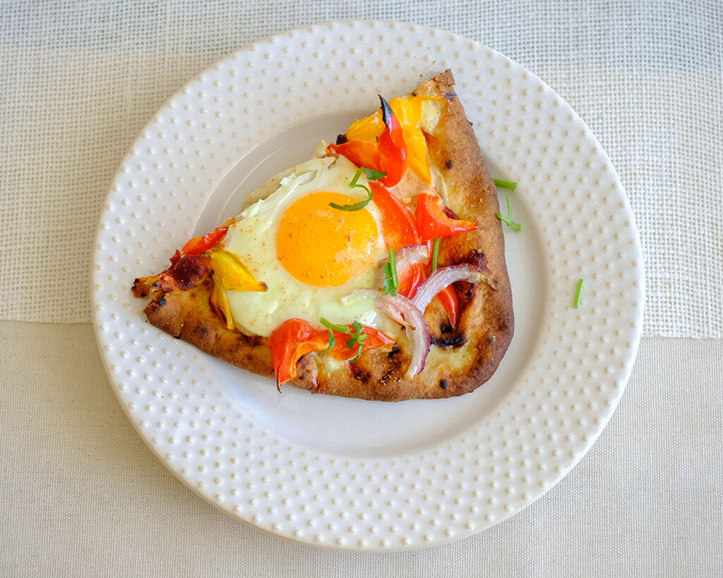 Round plate with breakfast pizza that has egg topping, bell pepper, and red onions.