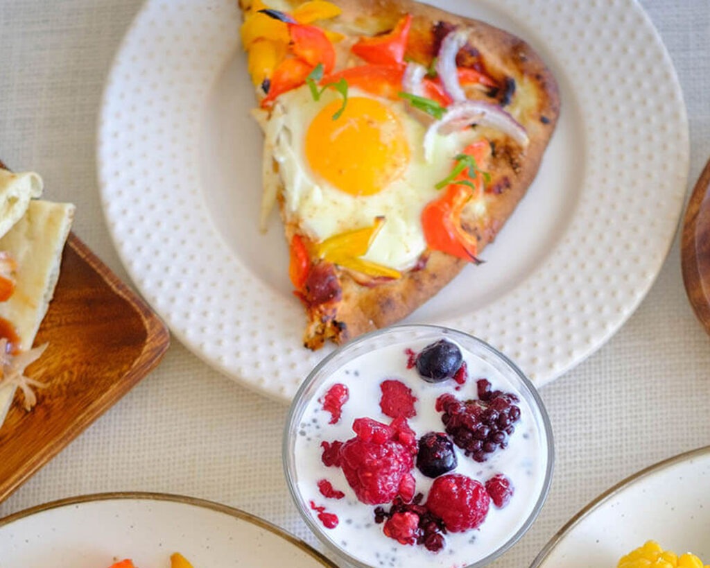 round plate with glass meal prep container with breakfast pizza with egg, naan bread, and vegetables