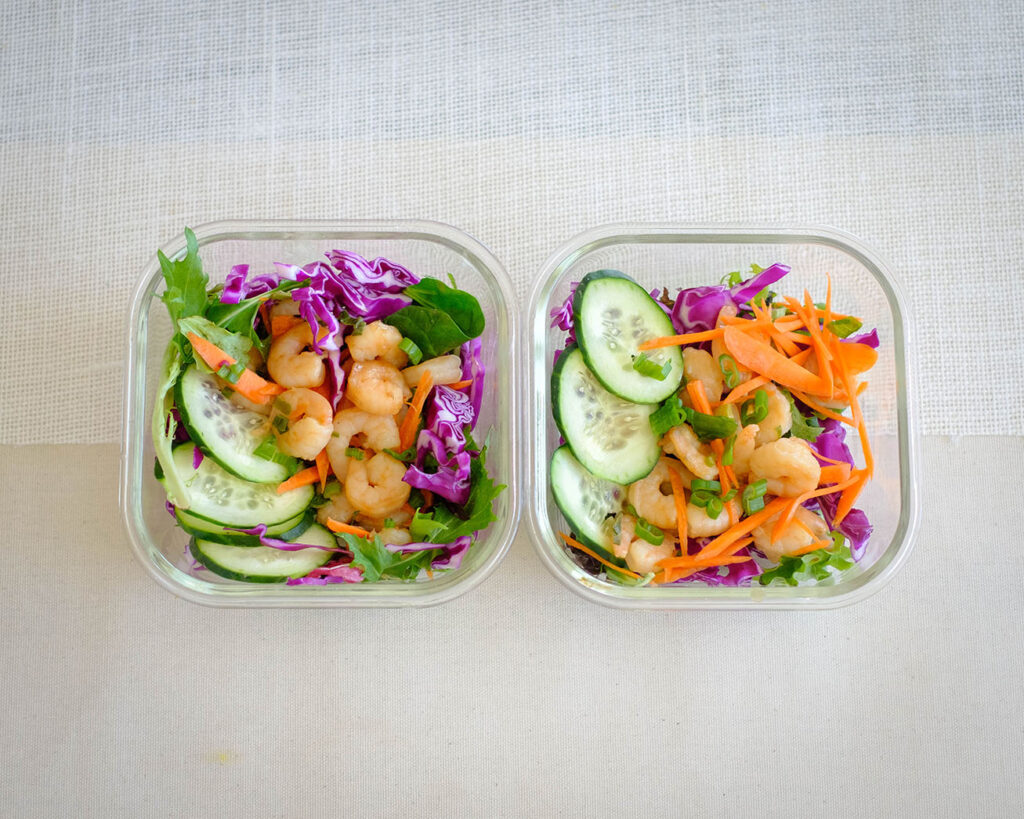 glass meal prep containers with salad, shrimp, and cucumbers, and carrots, and purple cabbage