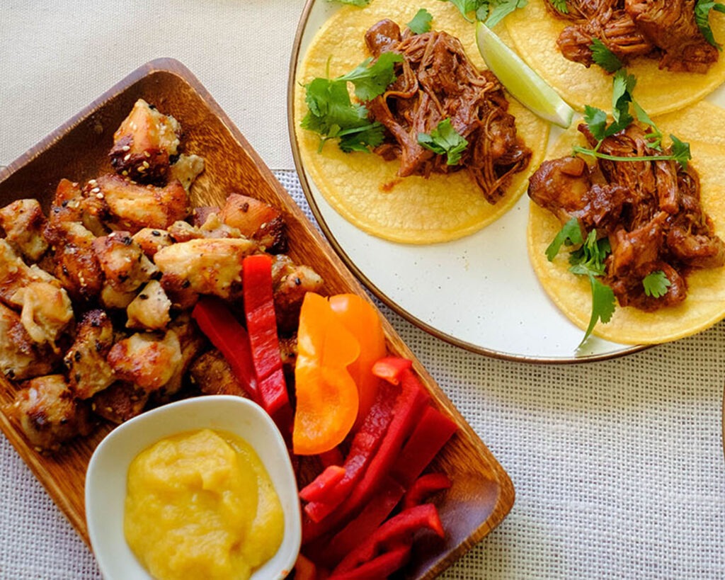 rectangular wooden plate with air fryer chicken recipe, small bite size chicken pieces, sliced red bell peppers, and honey mustard applesauce. Next to it is a round plate with chicken tacos