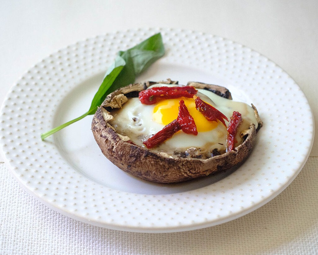 round plate with portobello mushroom baked with a egg and topped with sun-dried tomato slices and fresh basil.