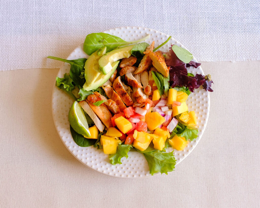 round plate with grilled chicken on top of lettuce with sliced avocado and mango salsa