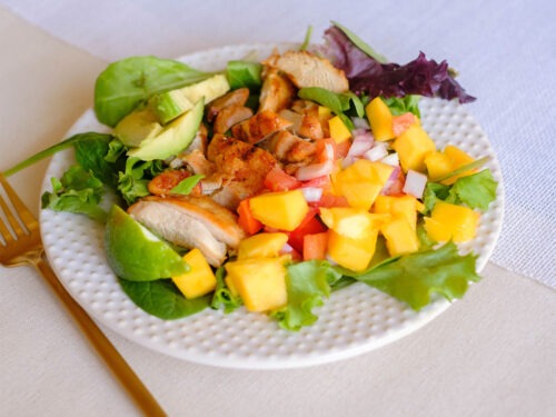 round plate with grilled chicken on top of lettuce with sliced avocado and mango salsa
