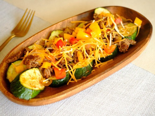 wooden plate with sliced zucchini topped with ground beef, bell peppers, tomatoes, and shredded cheese