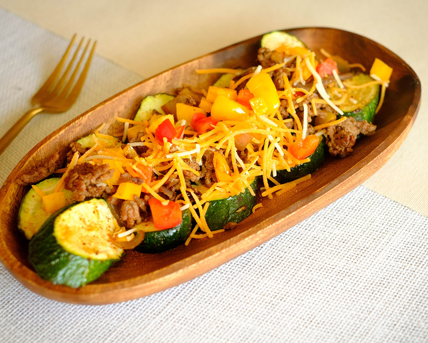 wooden plate with sliced zucchini topped with ground beef, bell peppers, tomatoes, and shredded cheese