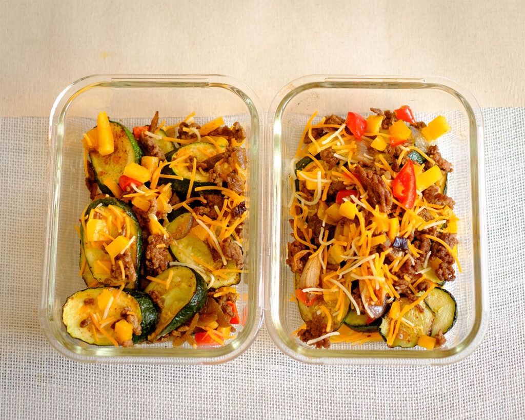 glass rectangular meal prep containers with sliced zucchini topped with ground beef, bell peppers, tomatoes, and shredded cheese