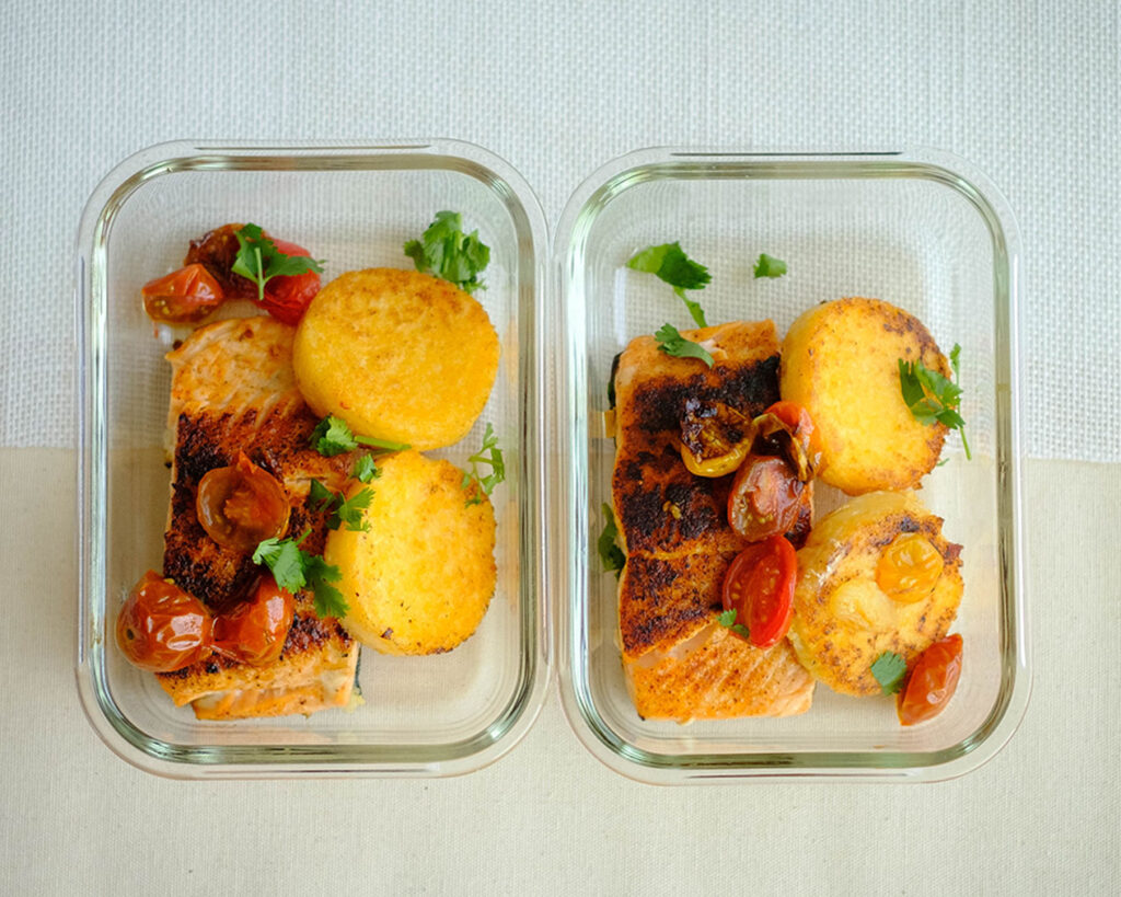 rectangular glass meal prep containers with salmon, cherry tomatoes, and polenta cakes