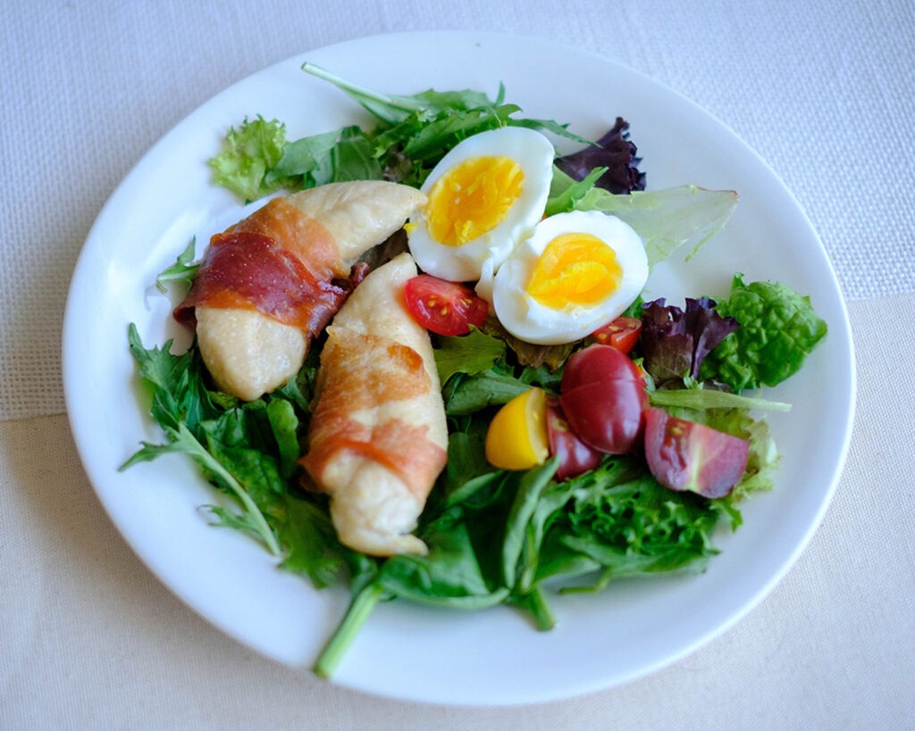 round plate with prosciutto wrapped chicken tenderloins, boiled eggs, cherry tomatoes, and spring mix salad