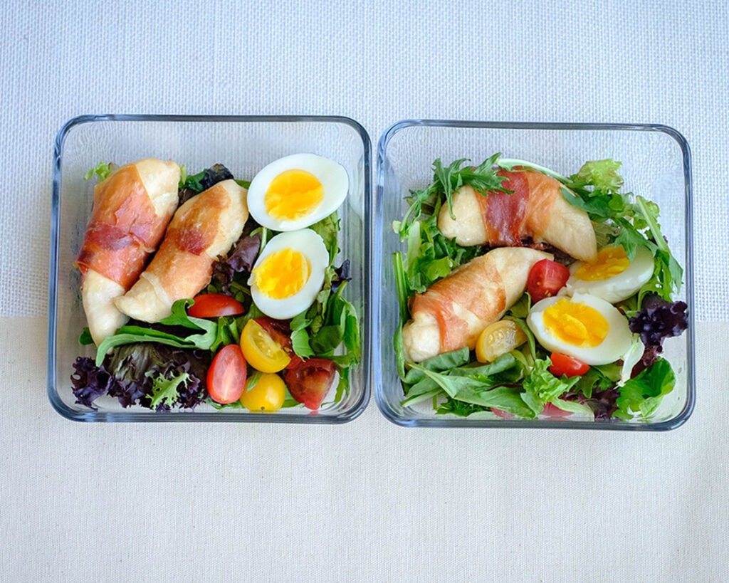 glass meal prep containers with prosciutto wrapped chicken tenderloins, boiled eggs, cherry tomatoes, and spring mix salad