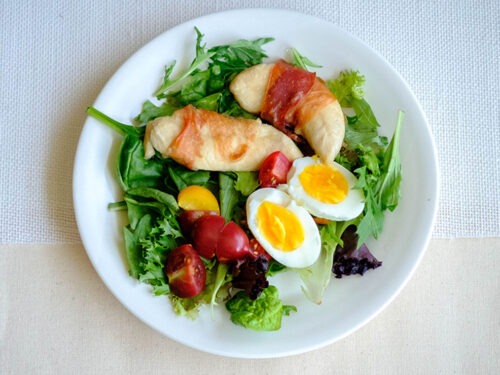 round plate with prosciutto wrapped chicken tenderloins, boiled eggs, cherry tomatoes, and spring mix salad