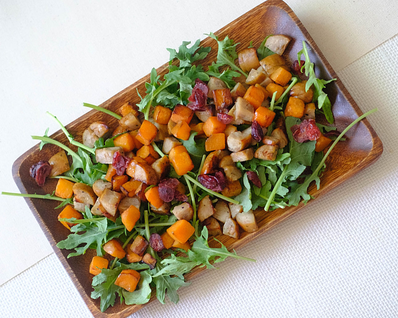 Rectangular wooden plate with diced sausage, diced butternut squash, dried cranberries, and green arugula salad.