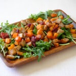 Rectangular wooden plate with diced sausage, diced butternut squash, dried cranberries, and green arugula salad.