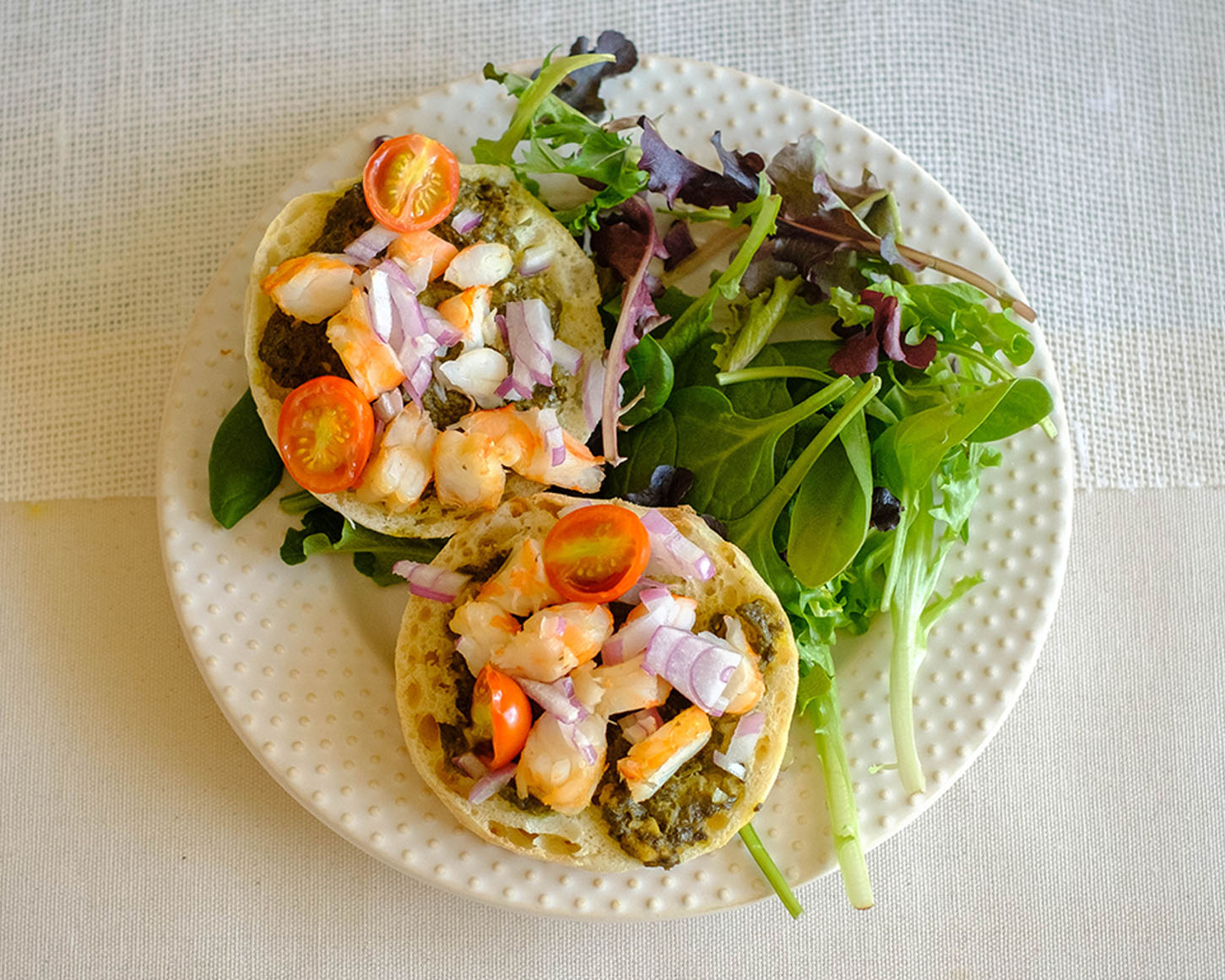 round plate with english muffins topped with chopped shrimp, red onions, and cherry tomatoes with spring mix salad