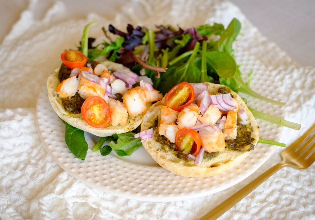 round plate with english muffins topped with chopped shrimp, red onions, and cherry tomatoes with spring mix salad