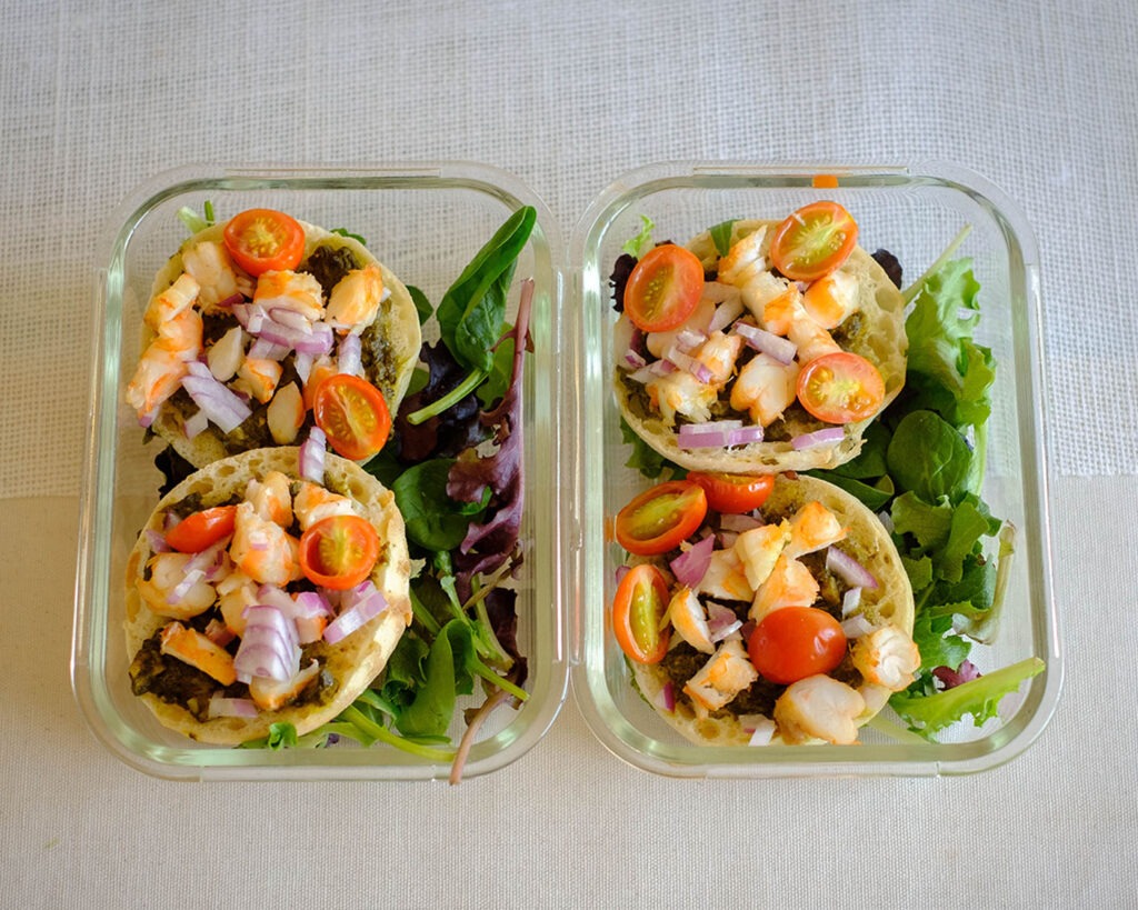 glass meal prep containers with english muffins topped with chopped shrimp, red onions, and cherry tomatoes with spring mix salad