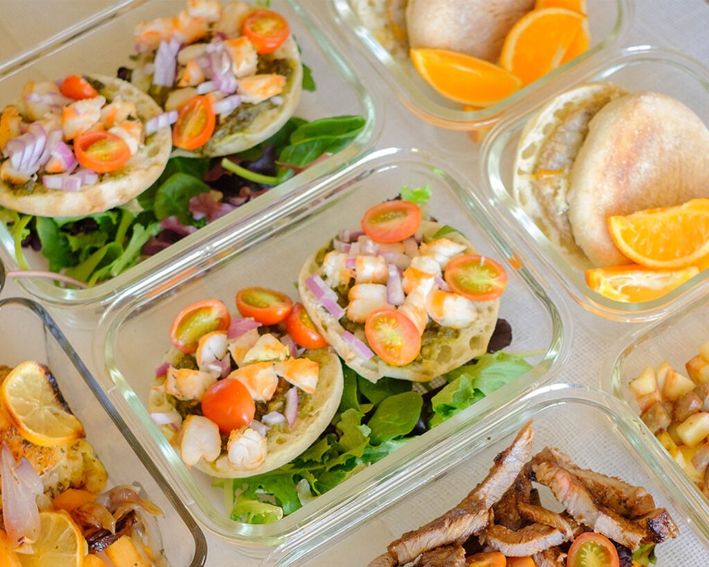 glass meal prep containers with english muffins topped with chopped shrimp, red onions, and cherry tomatoes with spring mix salad