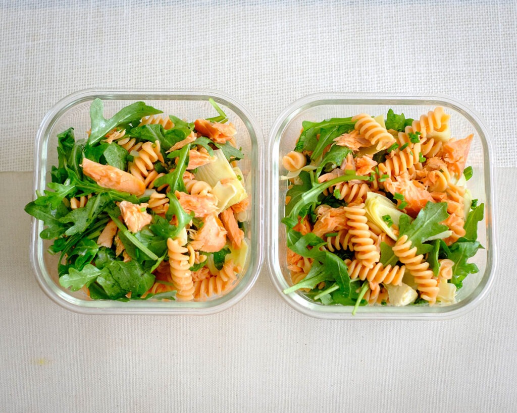 glass meal prep containers with rotini pasta, smoked salmon, artichoke hearts, and arugula