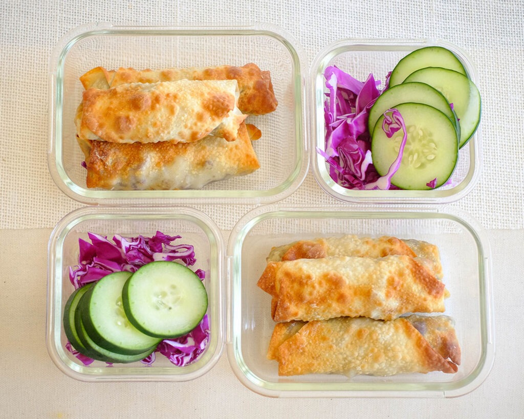 glass lunch meal prep containers with egg rolls, sliced green cucumber, and shredded red cabbage
