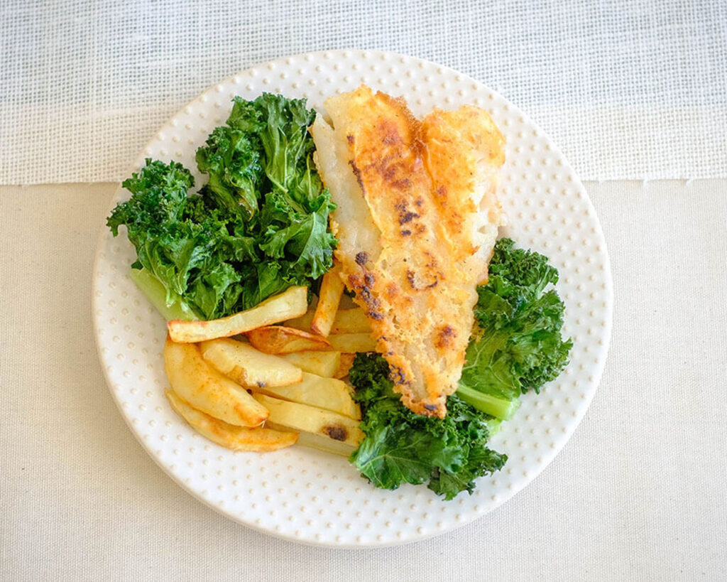 Round white plate with pan-fried fish, baked potato wedges and green kale
