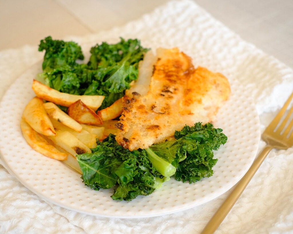 Round white plate with pan-fried fish, baked potato wedges and green kale