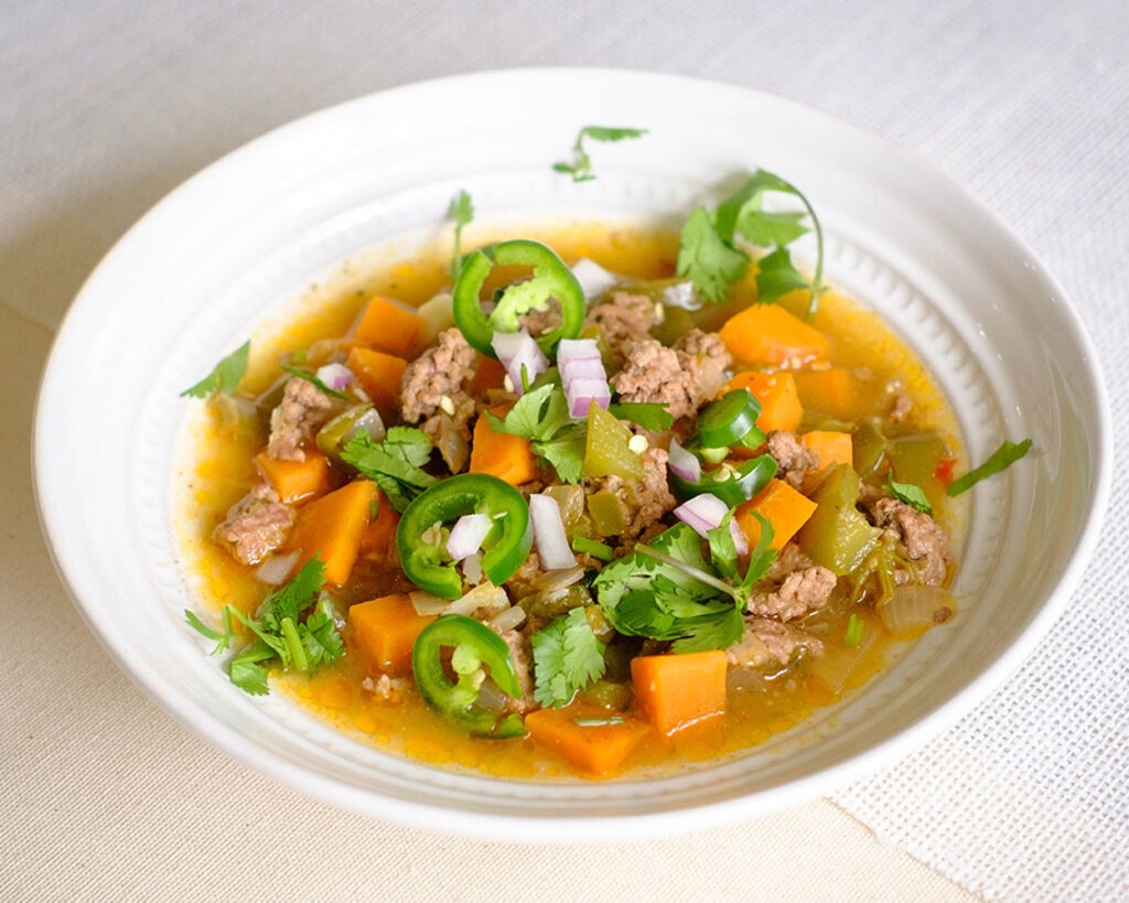 round bowl with soup that has ground beef, sweet potatoes, green bell peppers, cilantro, jalapeno, and red onions