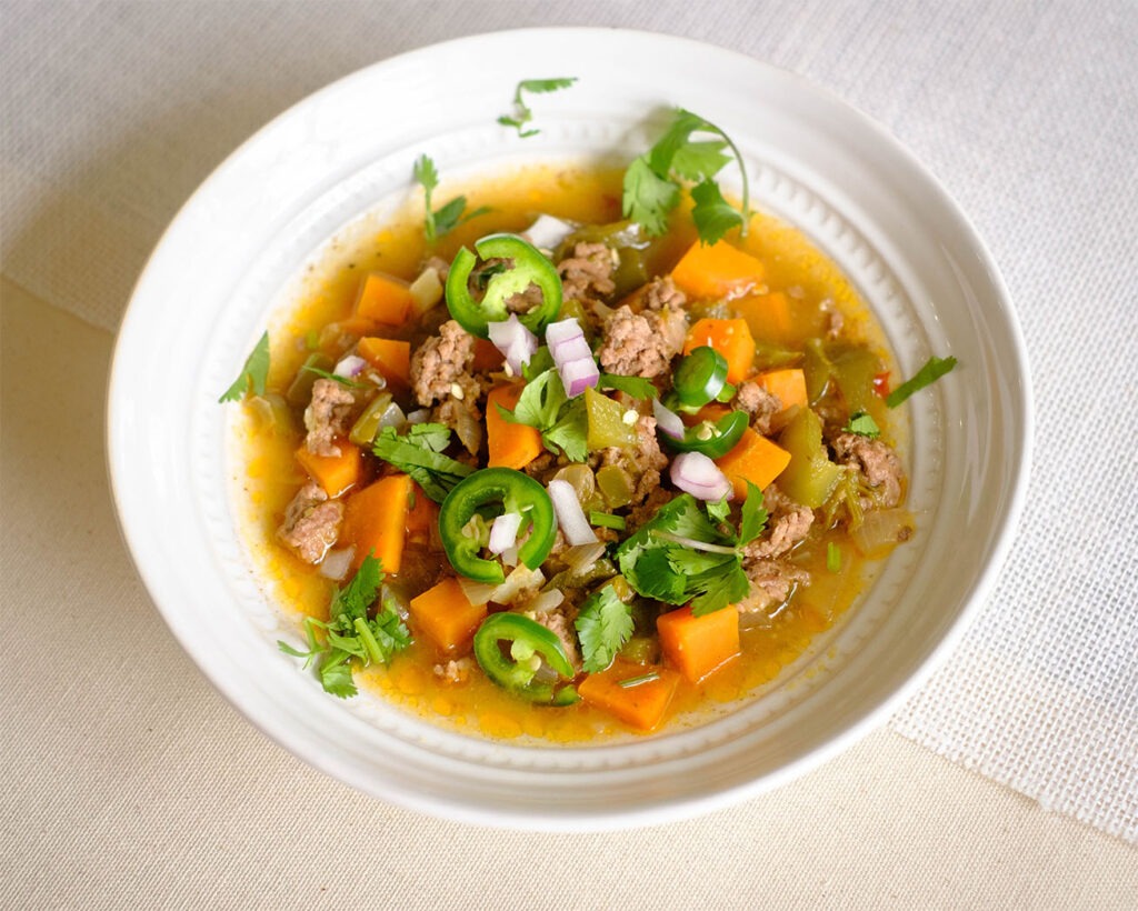 round bowl with soup that has ground beef, sweet potatoes, green bell peppers, cilantro, jalapeno, and red onions