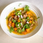 round bowl with soup that has ground beef, sweet potatoes, green bell peppers, cilantro, jalapeno, and red onions