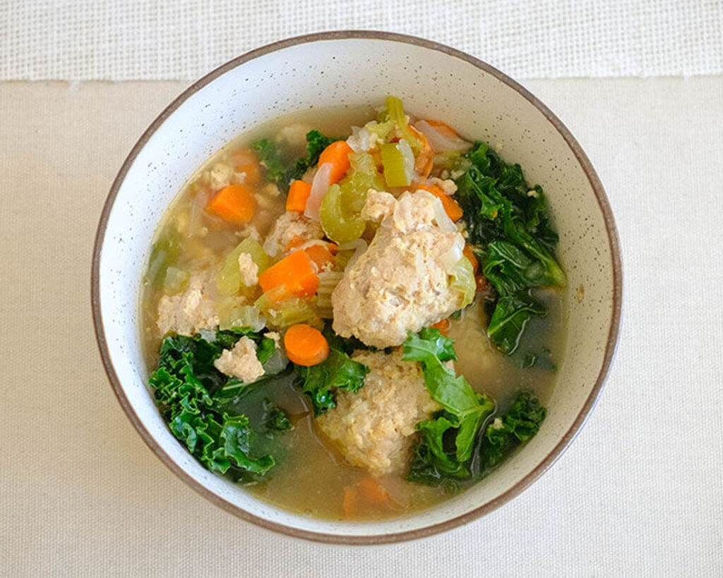 round bowl with chicken meatball soup, diced carrots, celery, onions, and baby kale