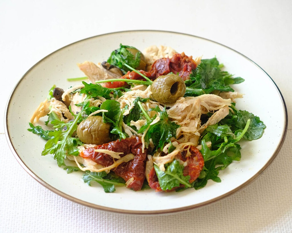 round plate with shredded chicken, sun-dried tomato, green olives, and baby kale