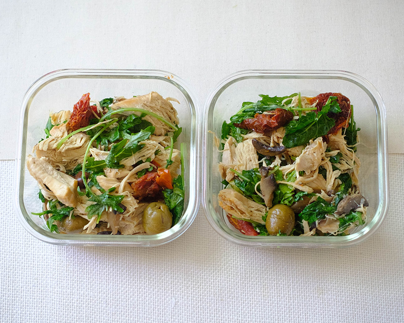 glass meal prep containers with shredded chicken, sun-dried tomato, green olives, and baby kale