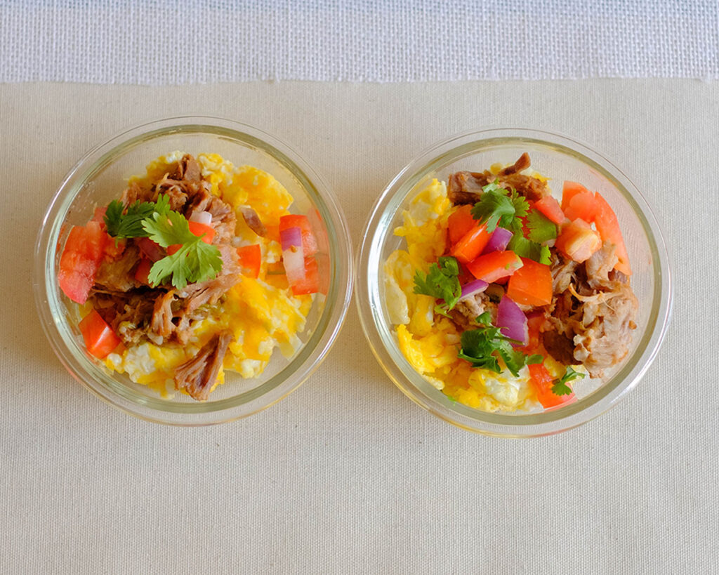 round glass meal prep containers with scrambled eggs, shredded pork, tomatoes, cilantro, and onions