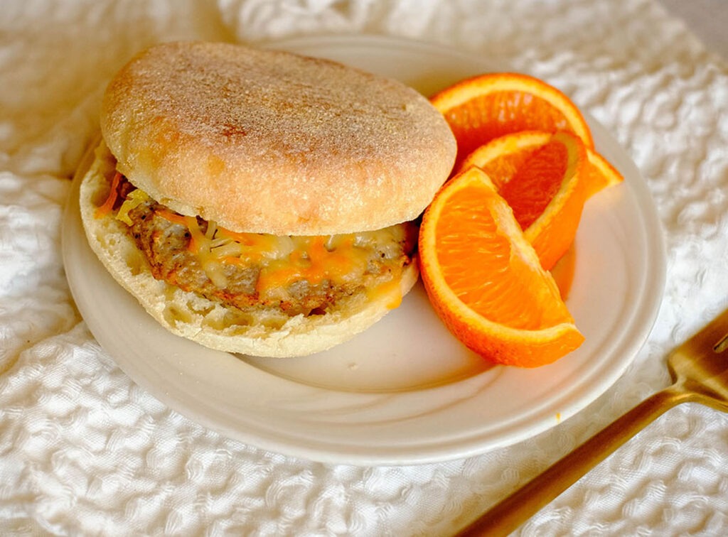 round plate with breakfast meal prep english muffin with a sausage patty, topped with shredded cheese and orange slices.