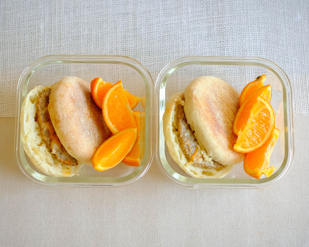 glass breakfast meal prep containers with english muffin with a sausage patty, topped with shredded cheese and orange slices.