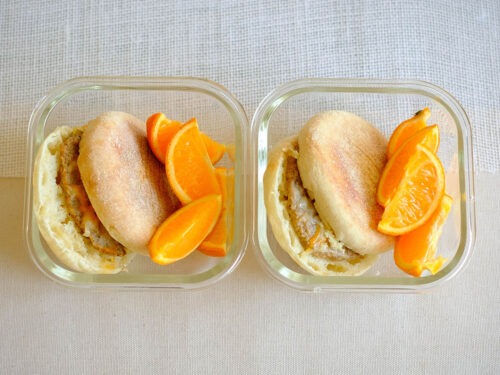 glass meal prep containers with english muffin with a sausage patty, topped with shredded cheese and orange slices.