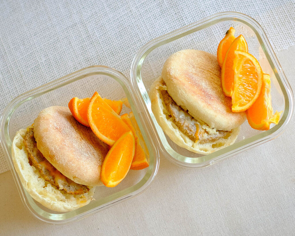 glass breakfast meal prep containers with english muffin with a sausage patty, topped with shredded cheese and orange slices.