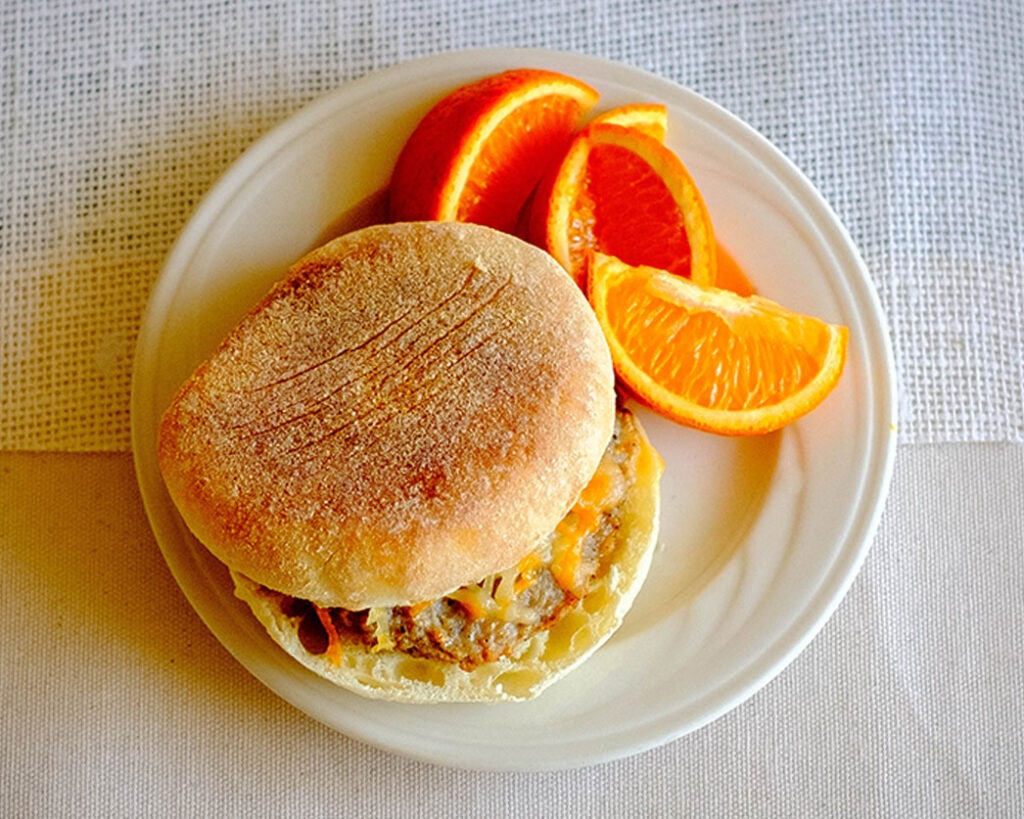 round plate with breakfast meal prep of english muffin with a sausage patty, topped with shredded cheese and orange slices.