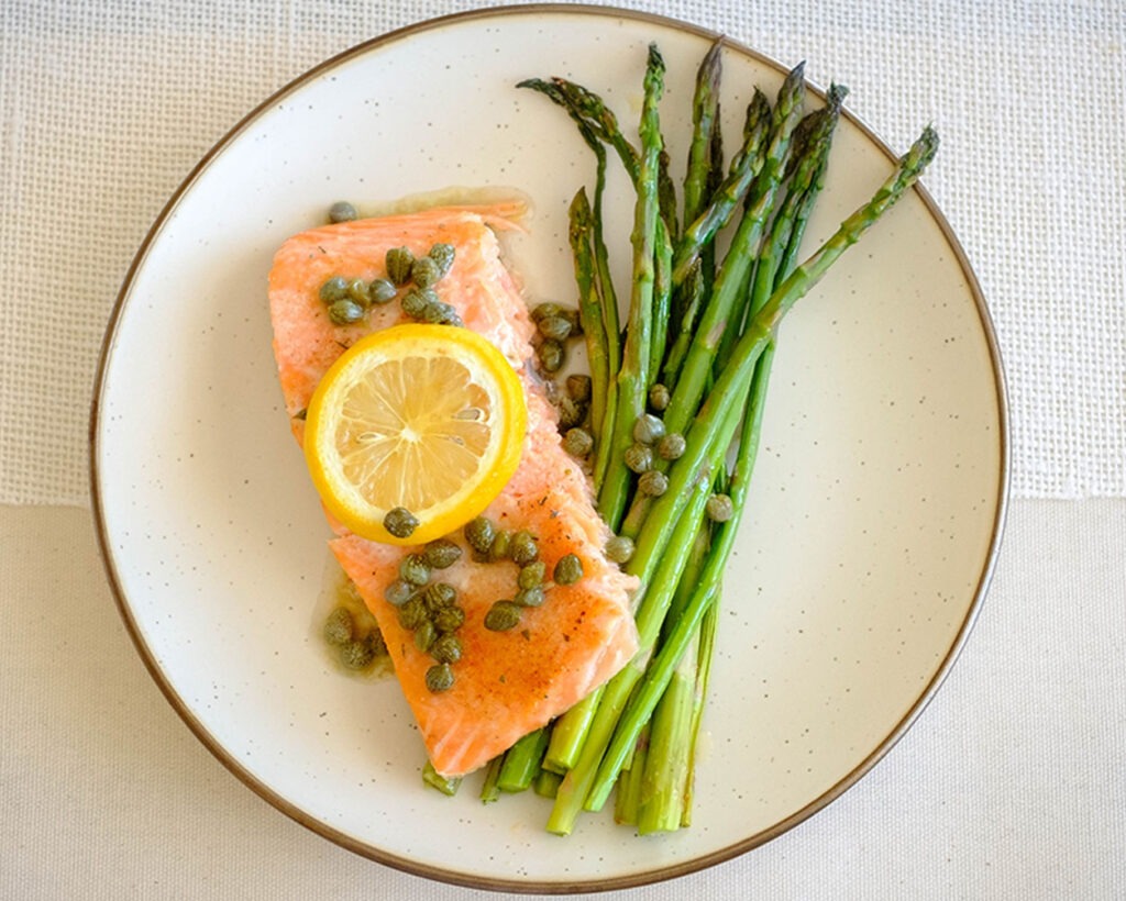 round plate with salmon fillet topped with lemon, capers, and asparagus