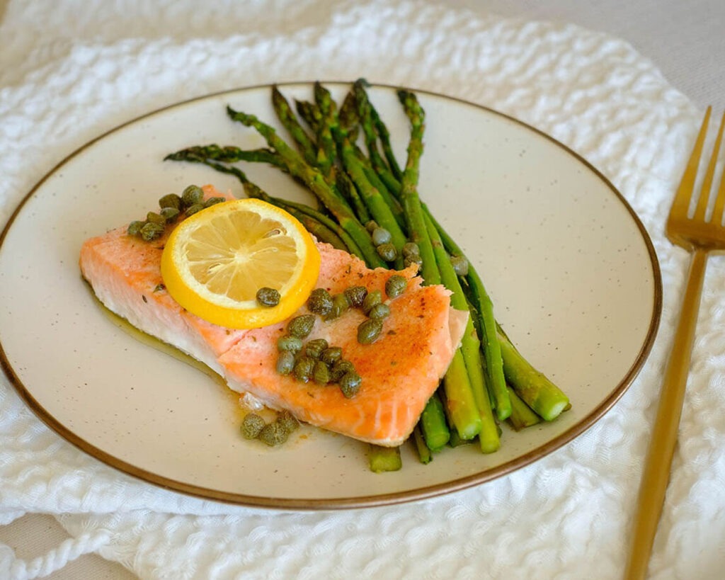 round plate with salmon fillet topped with lemon, capers, and asparagus