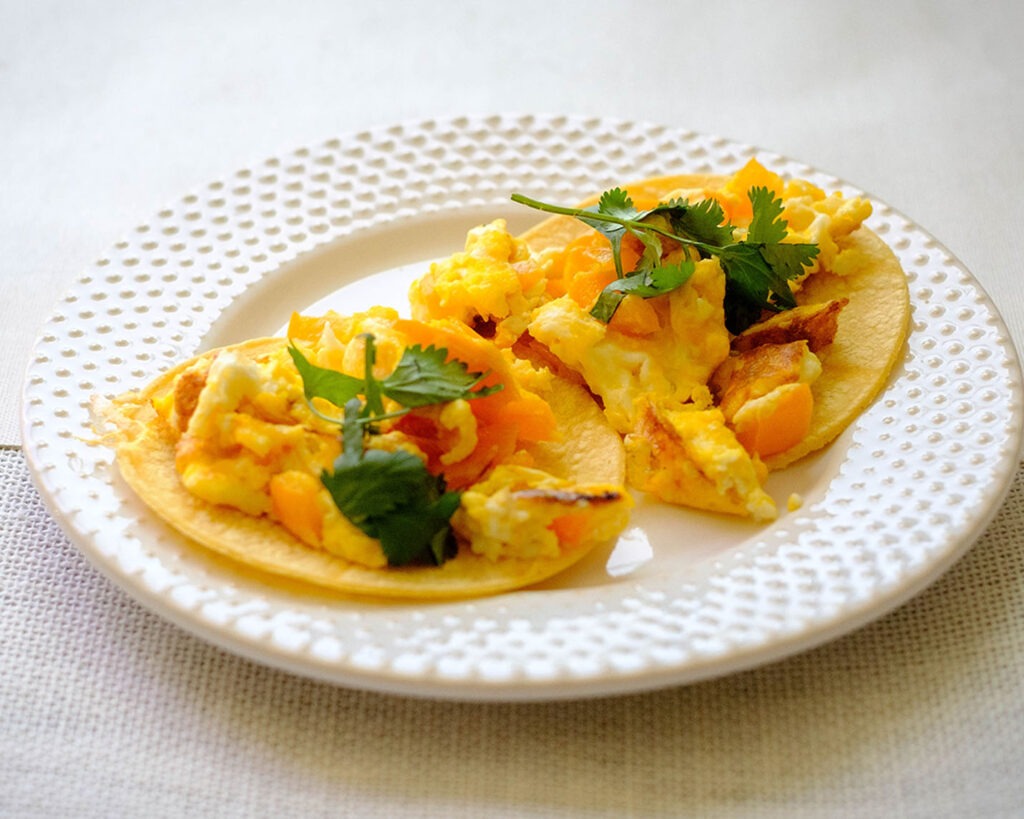 round plate with breakfast tacos made of corn tortillas topped with scrambled eggs and chopped cilantro and bell pepper