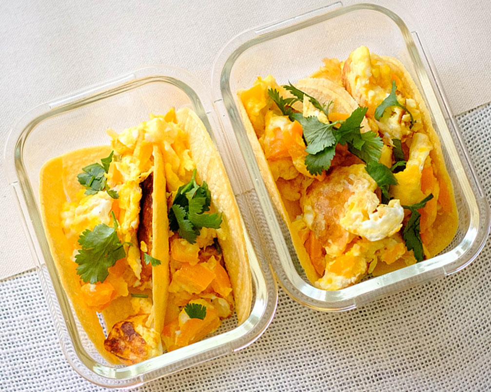 glass rectangular meal prep containers with breakfast tacos made of corn tortillas topped with scrambled eggs and chopped cilantro and bell pepper
